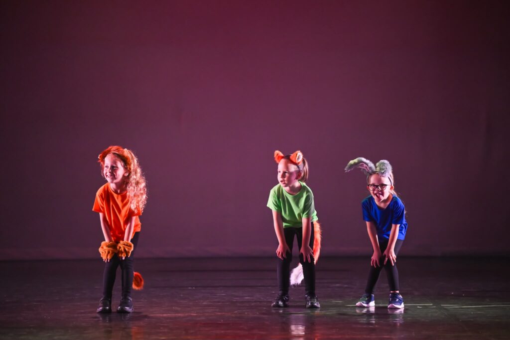 Four children in bright costumes with animal ears perform a stage dance, bending forward in a dark theater backdrop.