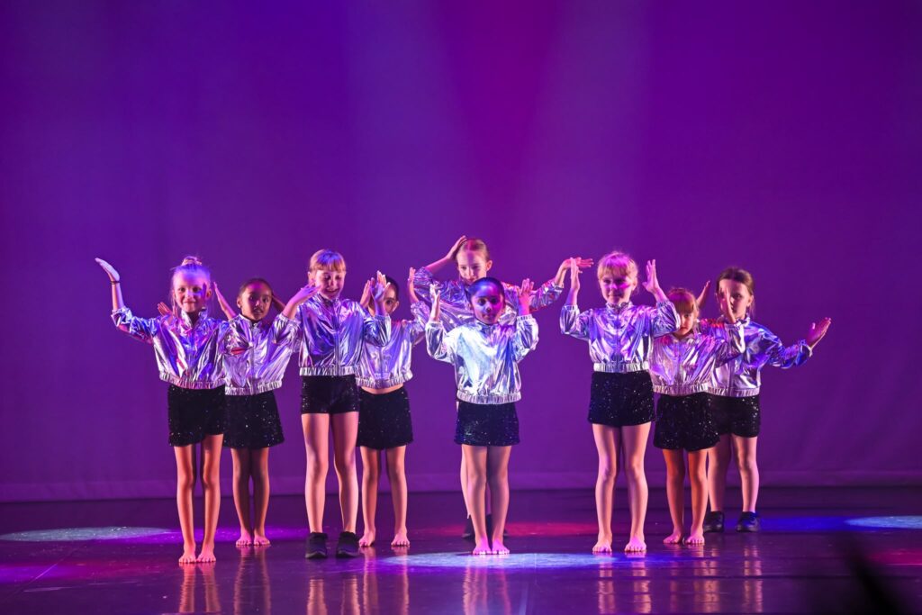 Group of young dancers on stage wearing shiny silver jackets and black skirts, arms raised under purple lights.