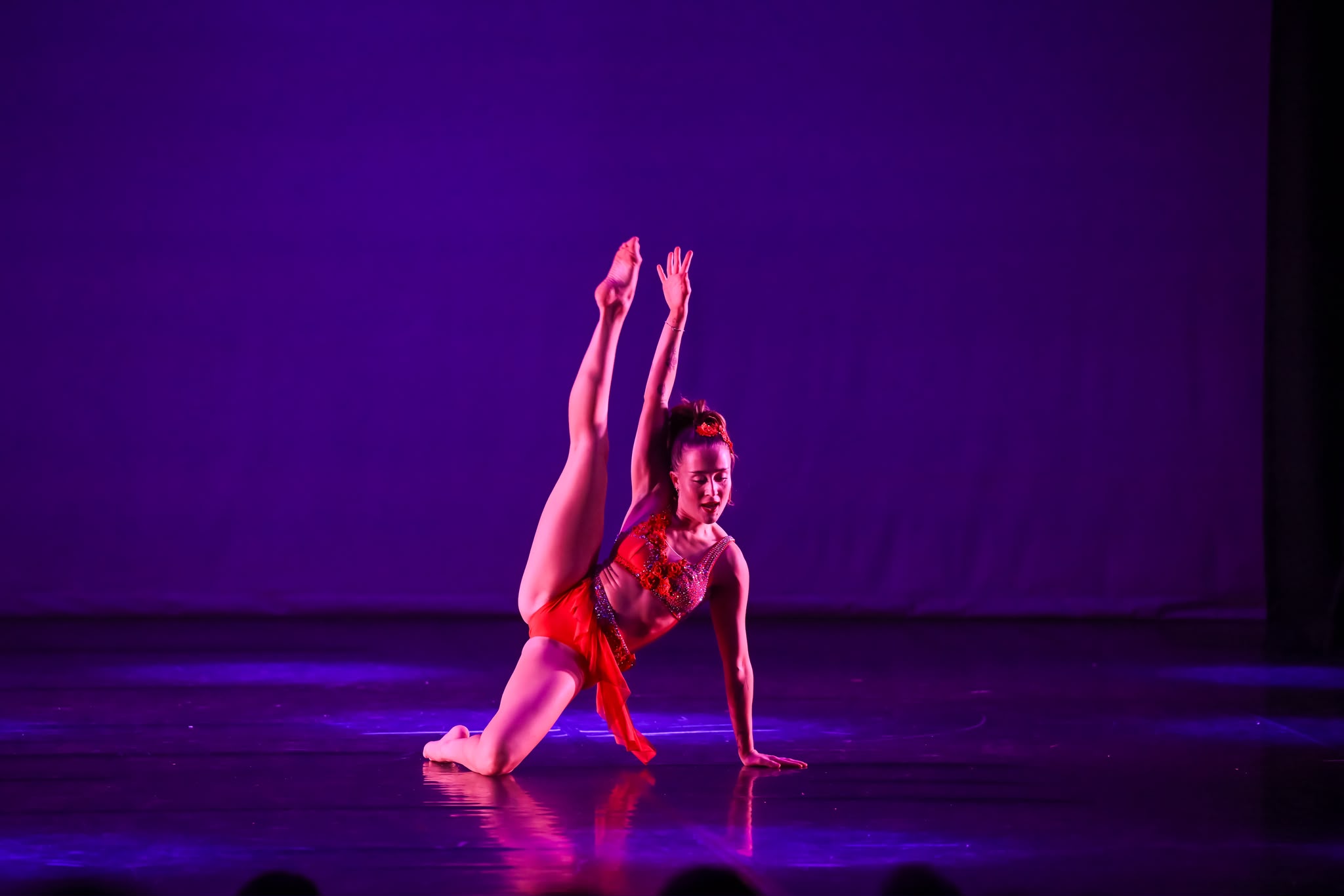 Female dancer in a red costume performs a dramatic pose on a dark purple-lit stage, one leg extended and arms raised above her head.
