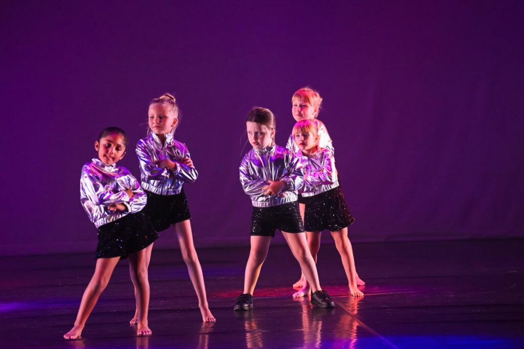Group of young girls in silver jackets and black sparkly skirts dancing on a dark purple-lit stage.